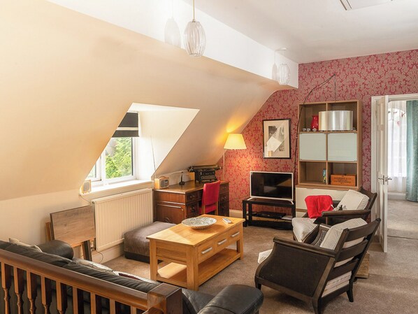 Ein gemütliches Dachzimmer mit schrägen Wänden, einem Holztisch, einem Fernseher, einem Bücherregal und einem Fenster mit Blick ins Grüne.