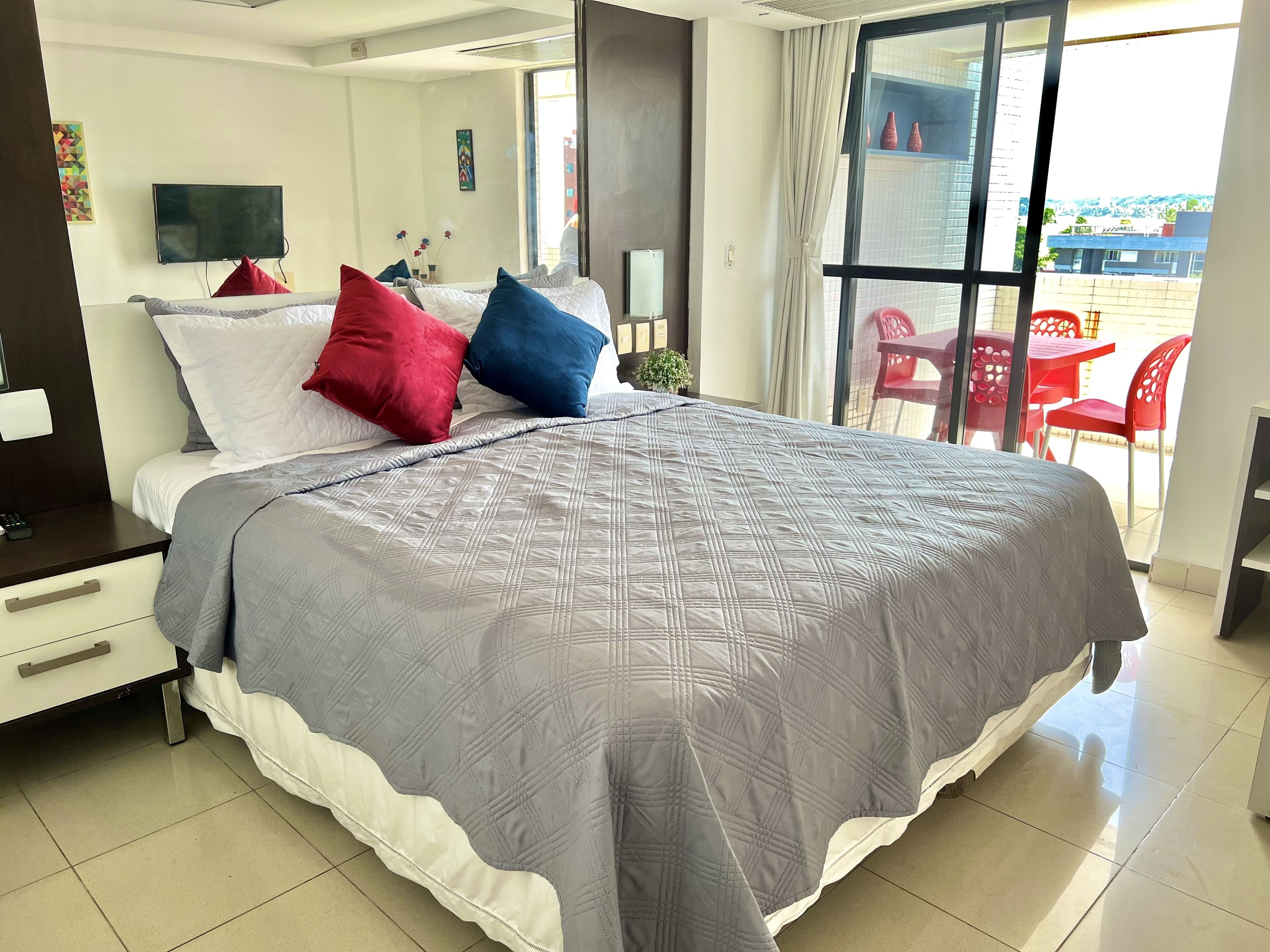 A modern hotel room with a large bed, a TV, a desk, and a balcony with red chairs.