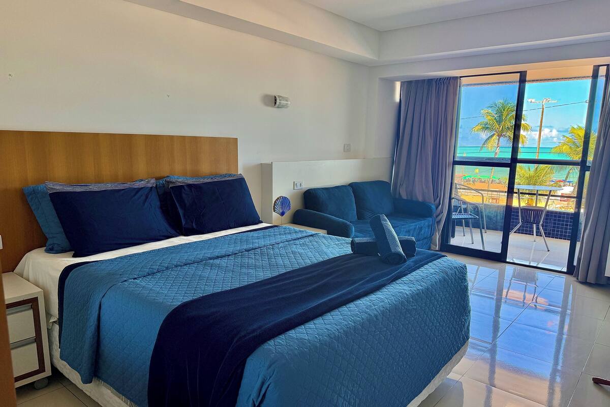 Ein Hotelzimmer mit einem großen Bett, einem Schreibtisch, einem Stuhl und einem Balkon mit Blick auf den Strand.