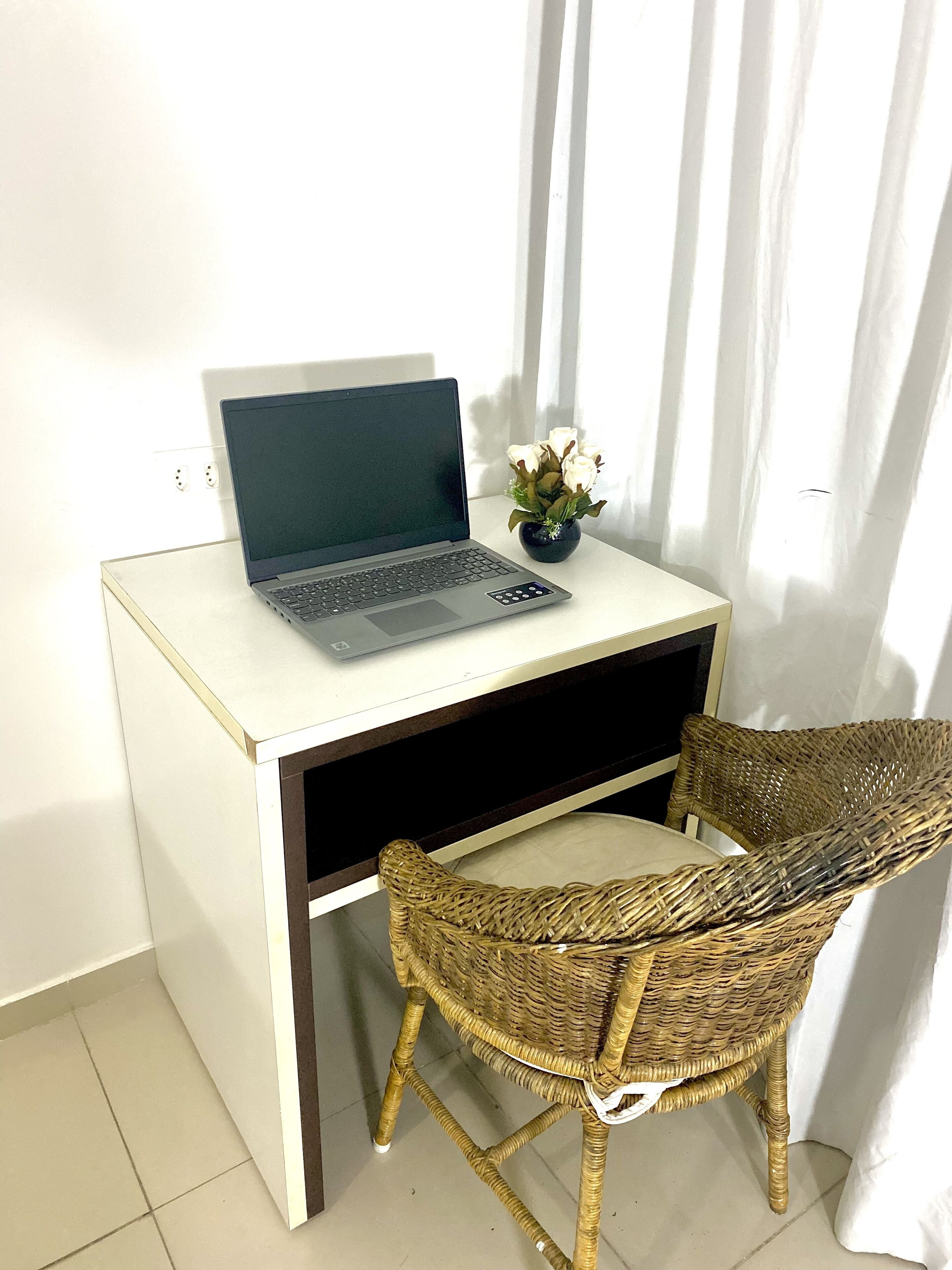 A small, white desk with a laptop and a vase of flowers. There is a wicker chair in front of the desk.
