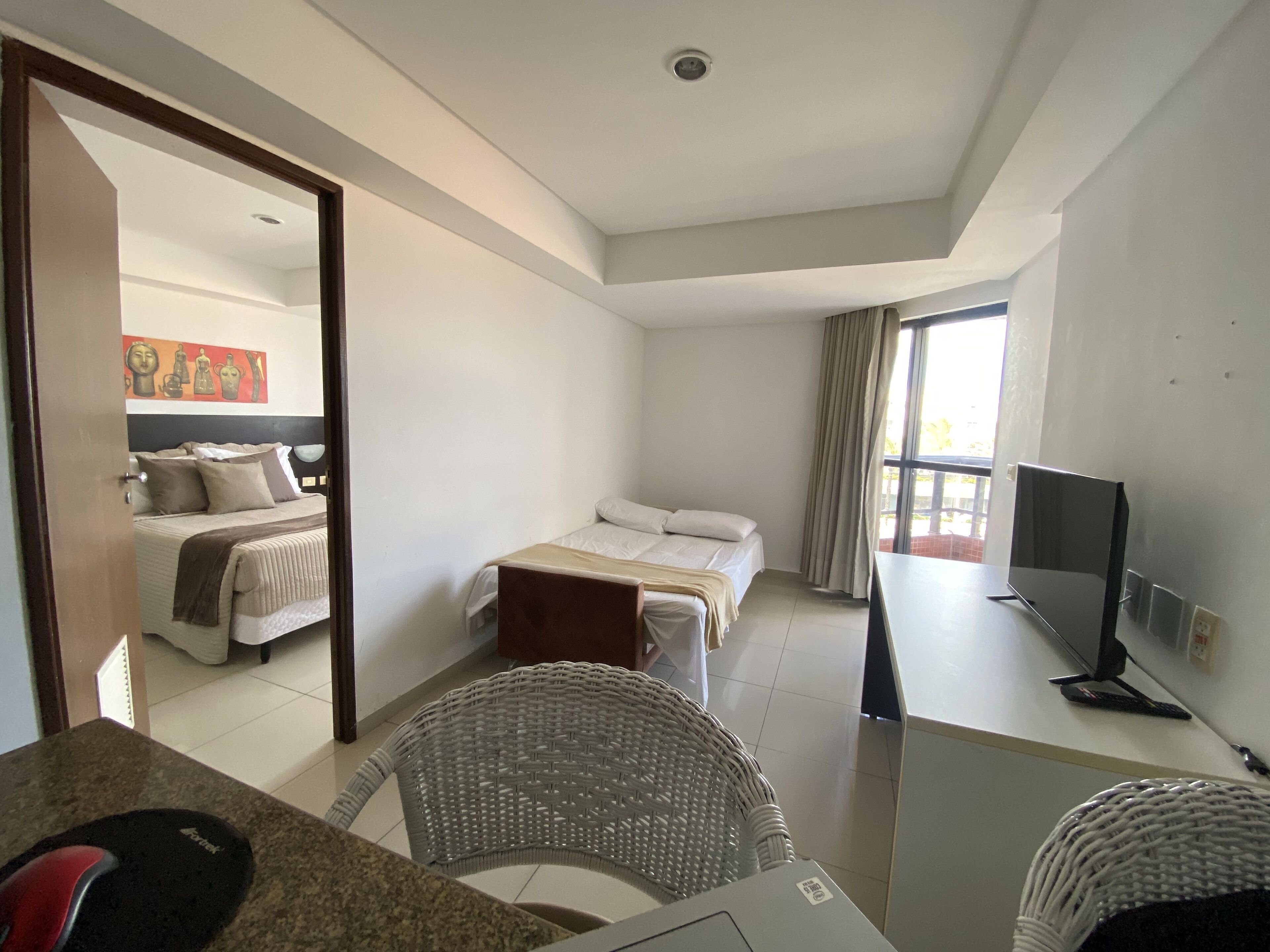 Quarto de hotel com cama, escrivaninha com computador, televisão e janela com cortinas.
