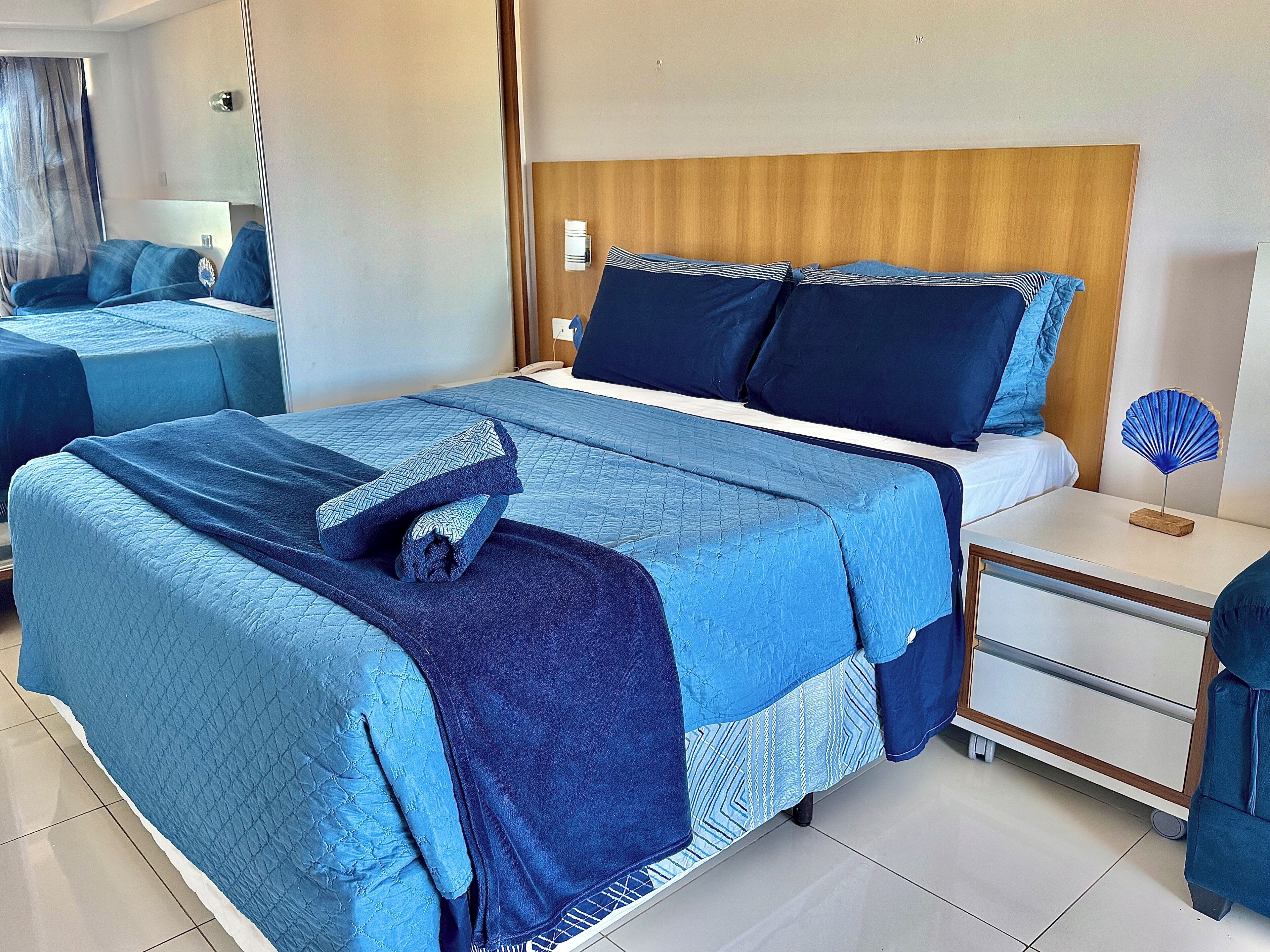 Quarto de hotel com uma cama grande, uma mesinha de cabeceira e um criado-mudo.