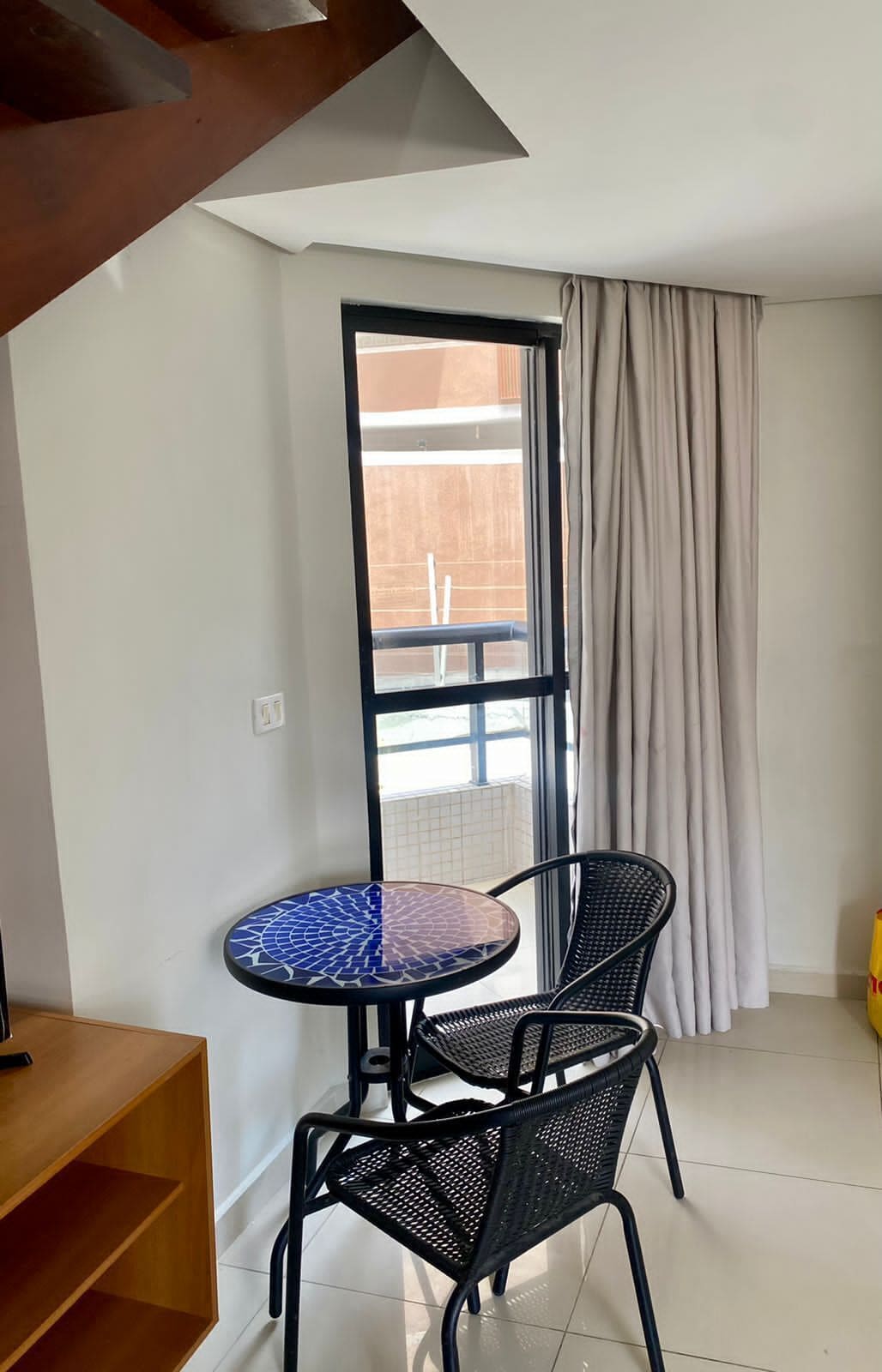 Um quarto pequeno com uma mesa redonda e duas cadeiras, uma prateleira de madeira e uma janela com cortinas.