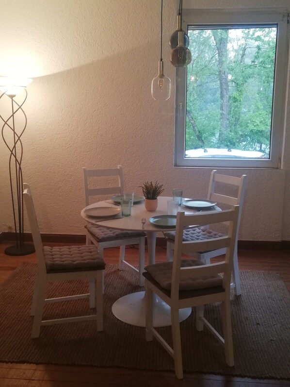 A dining area with a round table, four chairs, a potted plant, and a window with a view of trees.