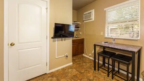 A compact kitchen with a white door, a built-in TV, a small table with stools, and a window with blinds.