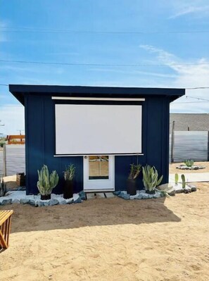 A small, dark blue building with a large white screen on the front, surrounded by potted plants and a sandy ground. - Sunburst House : Desert Chic Retreat (Joshua Tree)