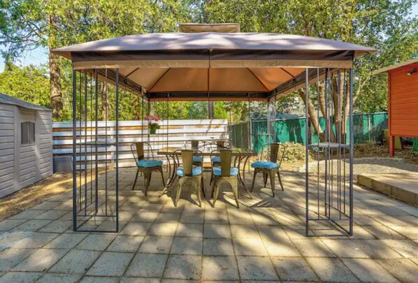 Relax under the shade of a covered seating area with a table and chairs, surrounded by a fence and a nearby building.