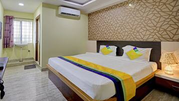 A hotel room with a bed, bedside lamp, air conditioning unit, and a window with curtains.