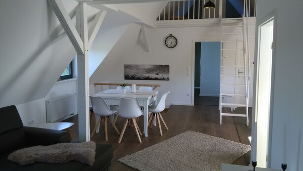 Una sala de estar moderna con un sofá oscuro, una mesa de comedor blanca con sillas, un cuadro en la pared y una escalera que sube a un nivel superior.