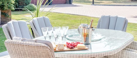 A round glass table with wicker chairs, a bottle of champagne, glasses, and a fruit platter.