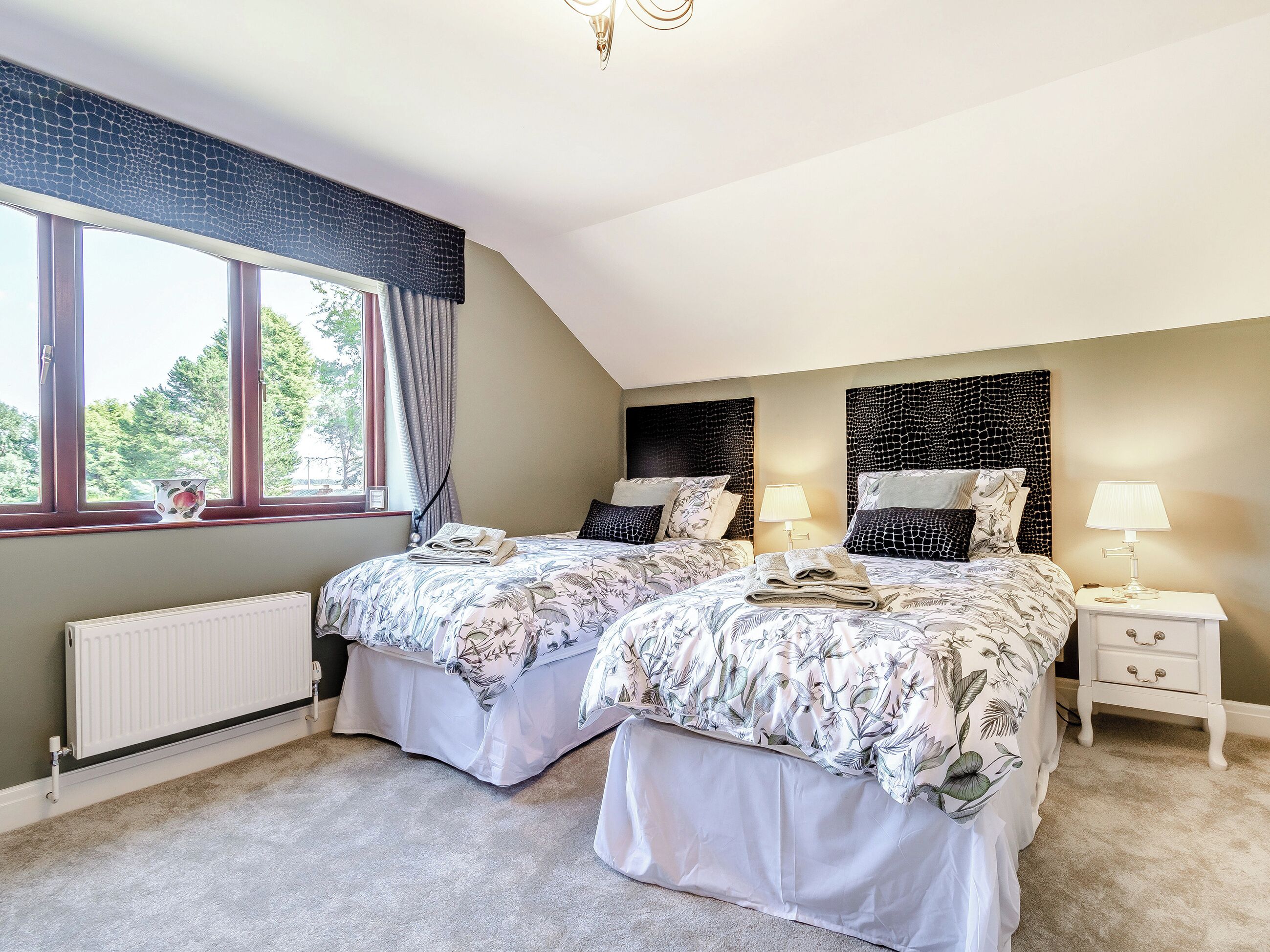 A bedroom with two beds, a window with curtains, a radiator, and a nightstand with a lamp.