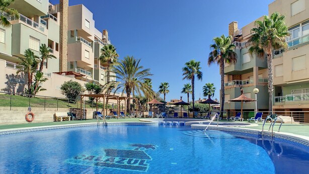 A swimming pool with a clear blue water feature, surrounded by palm trees and a multi-story building with balconies. - Apartment with mountain and sea views, parking (Cabo de Palos)
