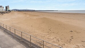 Una amplia playa arenosa con una baranda metálica en primer plano, una vista distante del mar y algunos edificios al fondo.
