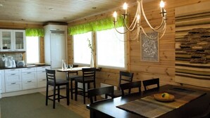Interior de una cabaña de madera con una mesa de comedor, sillas, una araña y una cocina con armarios y un refrigerador.