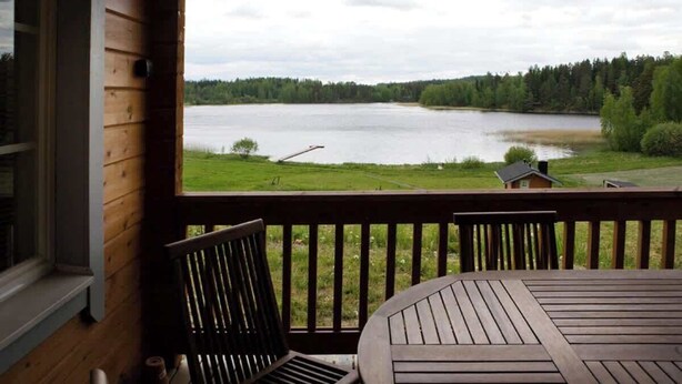 Terraza de madera con mesa y sillas, con vistas a un lago bordeado por un bosque.