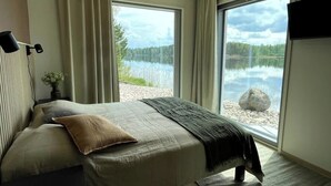 A bedroom with a bed, a bedside table, a lamp, and a view of a lake through large windows.