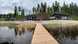 A wooden dock leading to a modern house with a dark exterior, surrounded by a natural landscape with trees and a body of water.