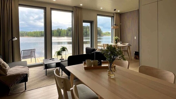 A modern living room with a large dining table, comfortable seating, and large windows overlooking a lake.