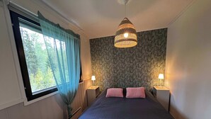 A bedroom with a bed, two bedside lamps, a window with curtains, a patterned wall, and a hanging light fixture.