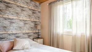 A bedroom with a wooden wall, a bed with a patterned bedspread, two pillows, a bedside table with a lamp, and sheer curtains on a window. - Enchanting cottage in the woods, by the lake (Orivesi)