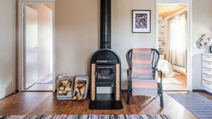 A room with a wood-burning stove, a chair, a bed, and a door leading to another room. - Enchanting cottage in the woods, by the lake (Orivesi)