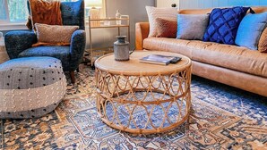 Een gezellige woonkamer met een kamerkleed met patroon, een ronde rieten salontafel, een bruine leren bank en een blauwe fauteuil.