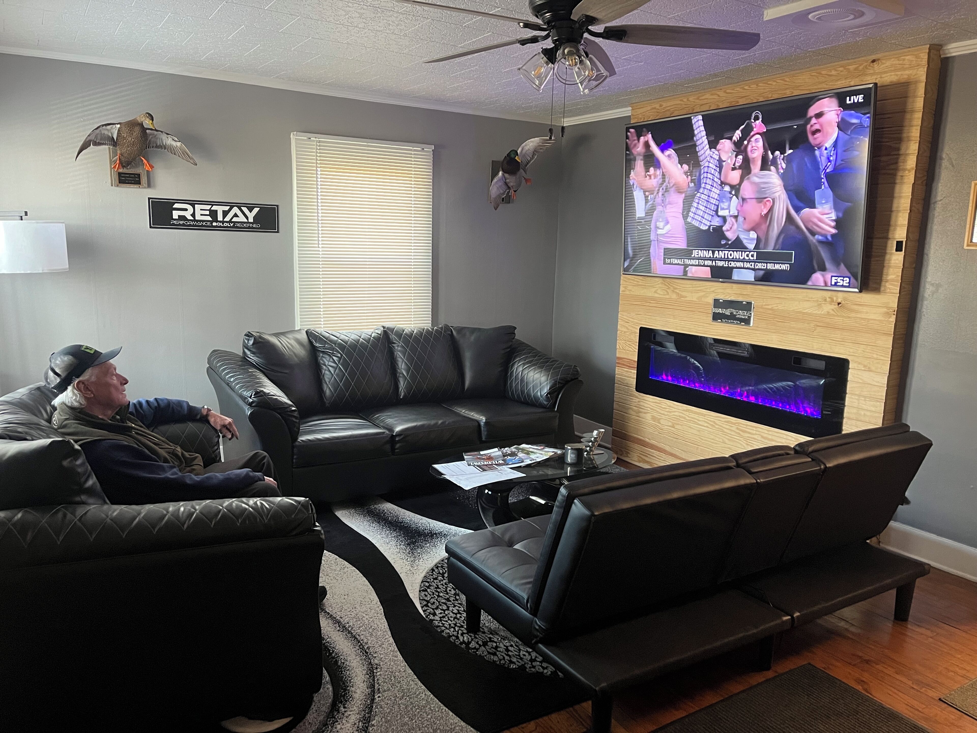 A living room with a large flat-screen TV mounted on a wooden wall, leather sofas, a ceiling fan, and a 'RETAIL' sign on the wall.