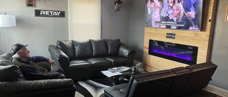 A living room with a large flat-screen TV mounted on a wooden wall, leather sofas, a ceiling fan, and a 'RETAIL' sign on the wall.