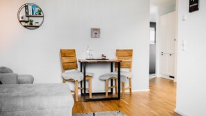 Ein moderner, hell eingerichteter Raum mit einem grauen Sofa, einem kleinen Tisch mit zwei Stühlen, einem Bücherregal und einem gerahmten Bild an der Wand.