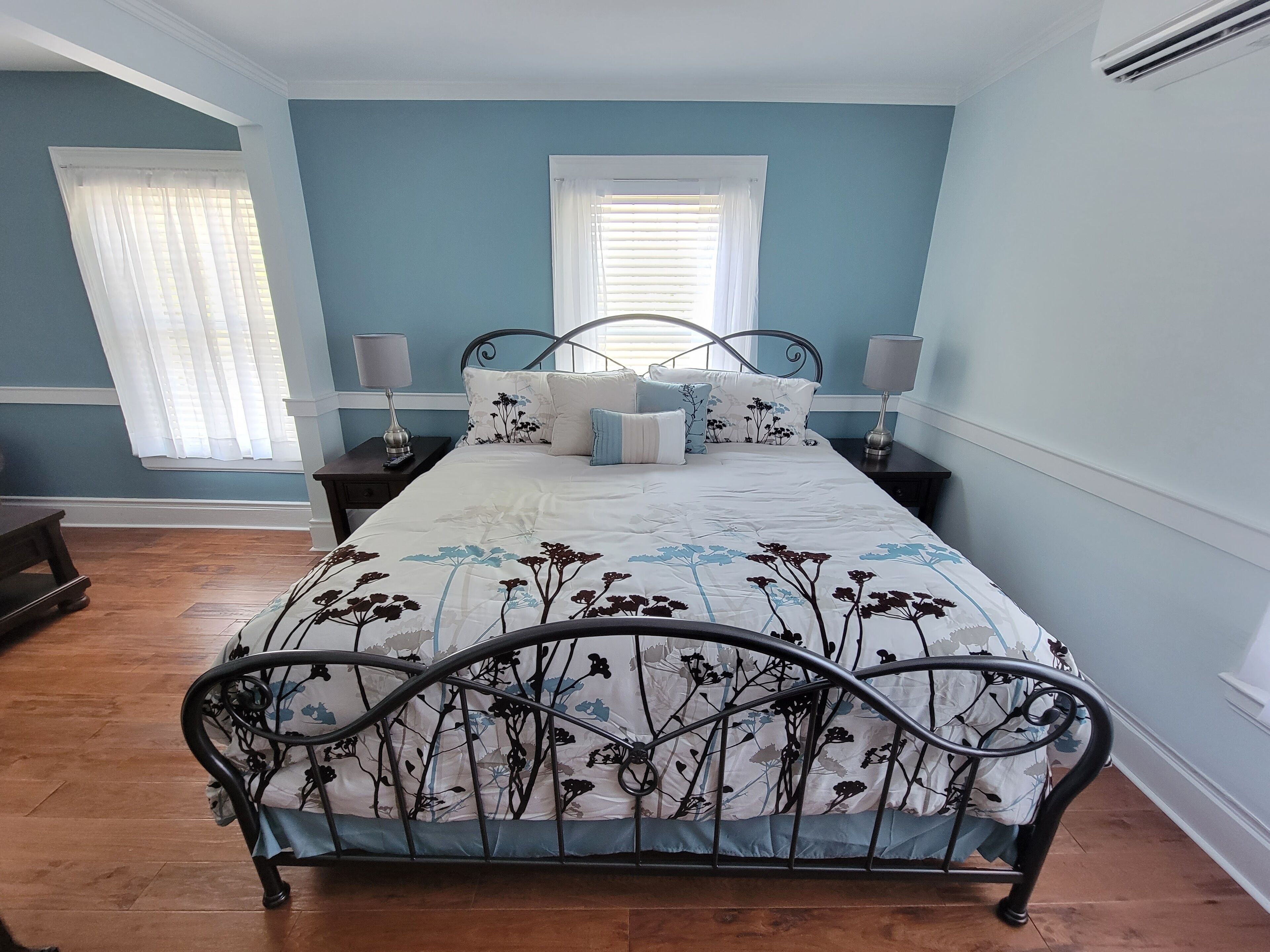 A bedroom with a bed, two bedside tables, a window with blinds, and a wooden floor.