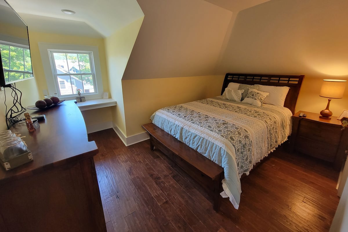A bedroom with a bed, bedside table, lamp, and a window with a view of greenery.