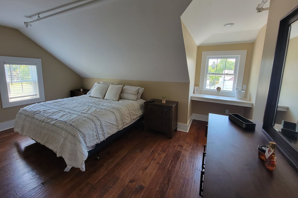 A bedroom with a sloped ceiling, a bed with a white quilt, a nightstand, a dresser, and a window with blinds.
