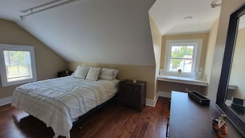 Ein Schlafzimmer mit schrägen Wänden, einem Bett mit weißer Decke, einem Nachttisch, einer Kommode und einem Fenster mit Jalousien.