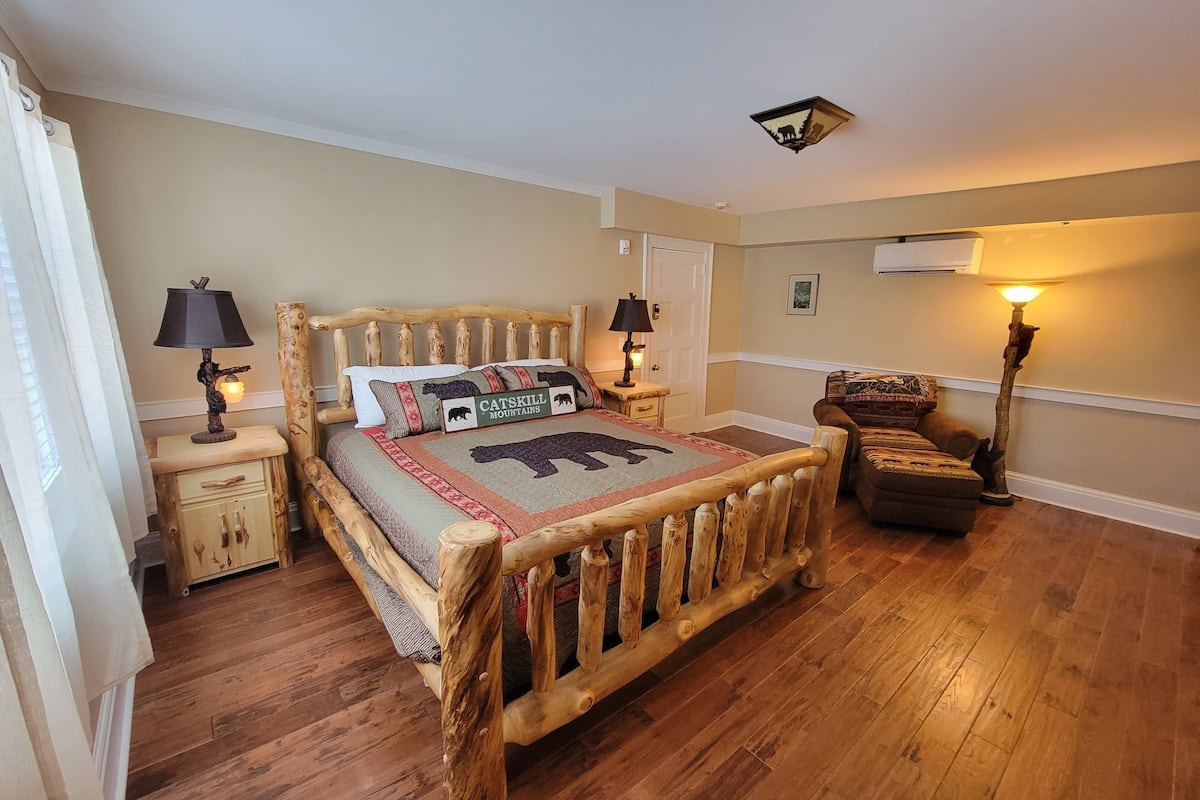 A cozy bedroom with a wooden bed, two bedside tables with lamps, a chair, and a large window with curtains.