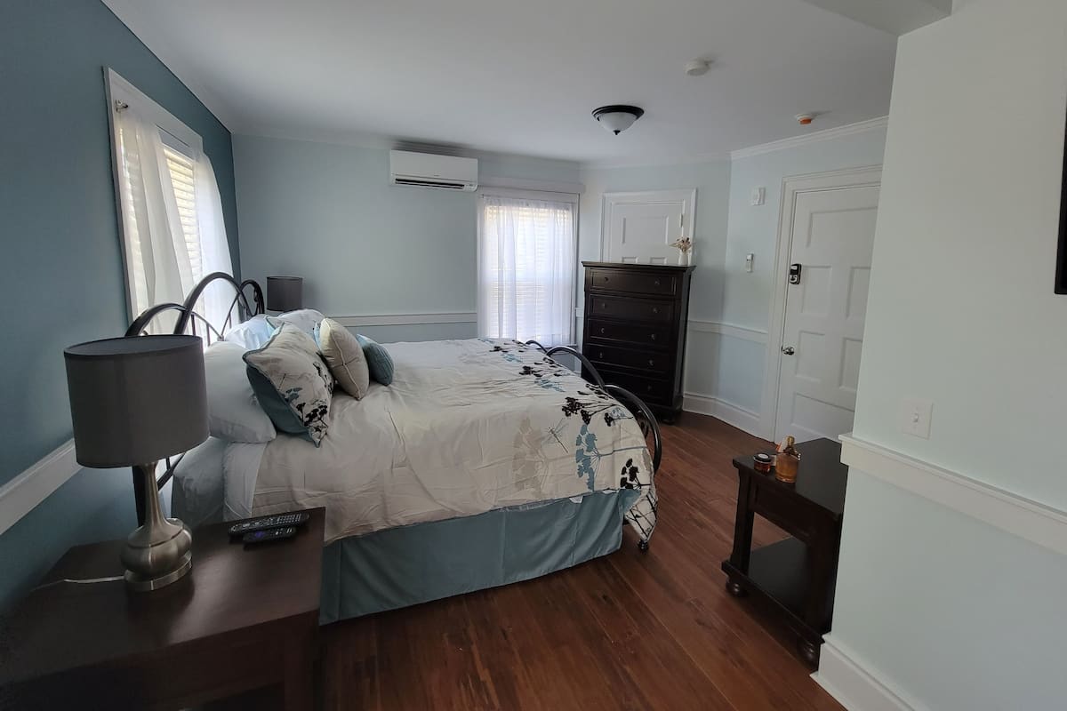A bedroom with a bed, bedside table, lamp, dresser, and an air conditioning unit.