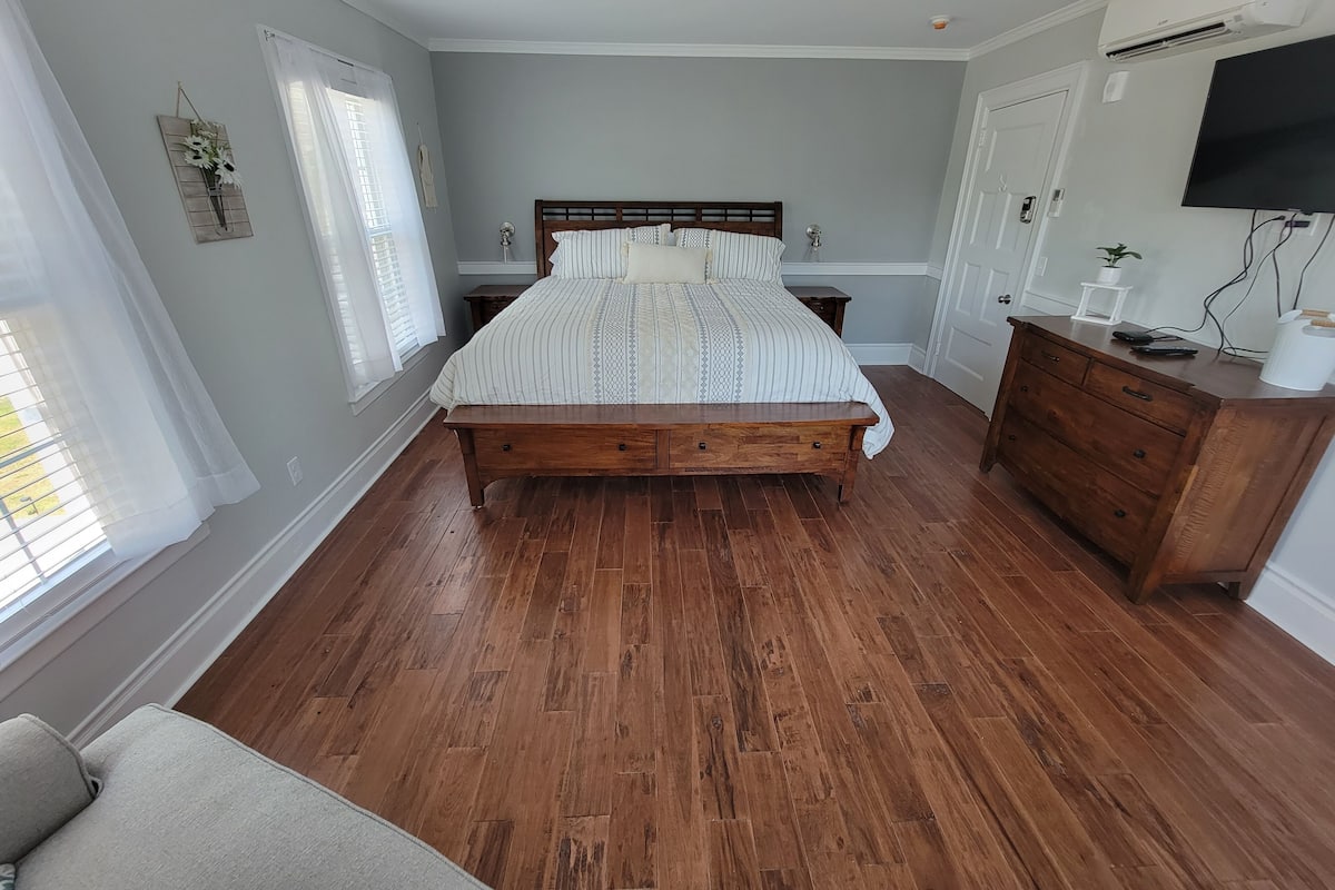 A bedroom with a wooden bed, a dresser, a TV, and a window with sheer curtains.