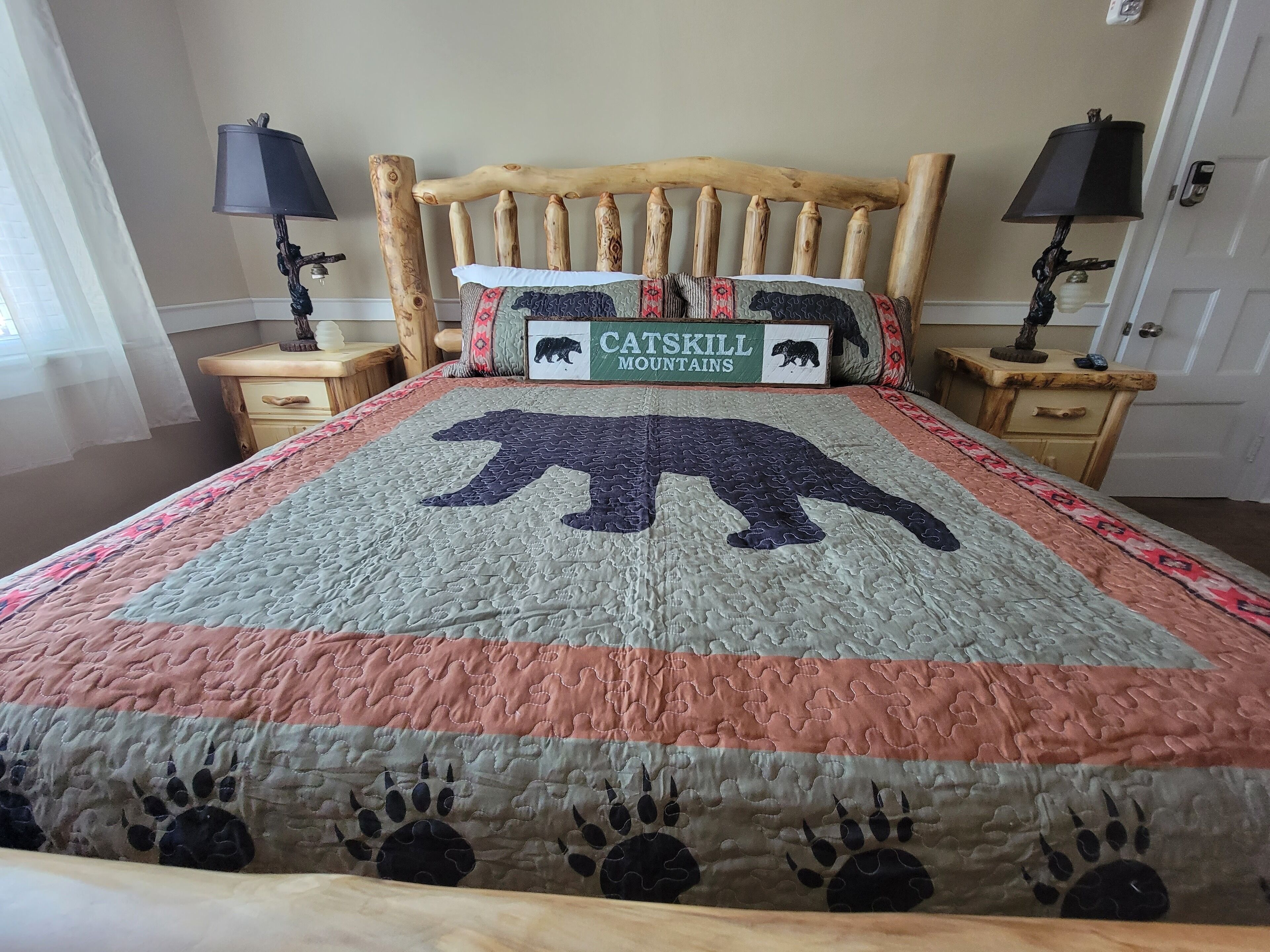 Una cama con colcha que presenta un oso y huellas de pata, cabecera de madera, dos mesitas de noche con lámparas y un cartel que dice "Catskill Mountains".