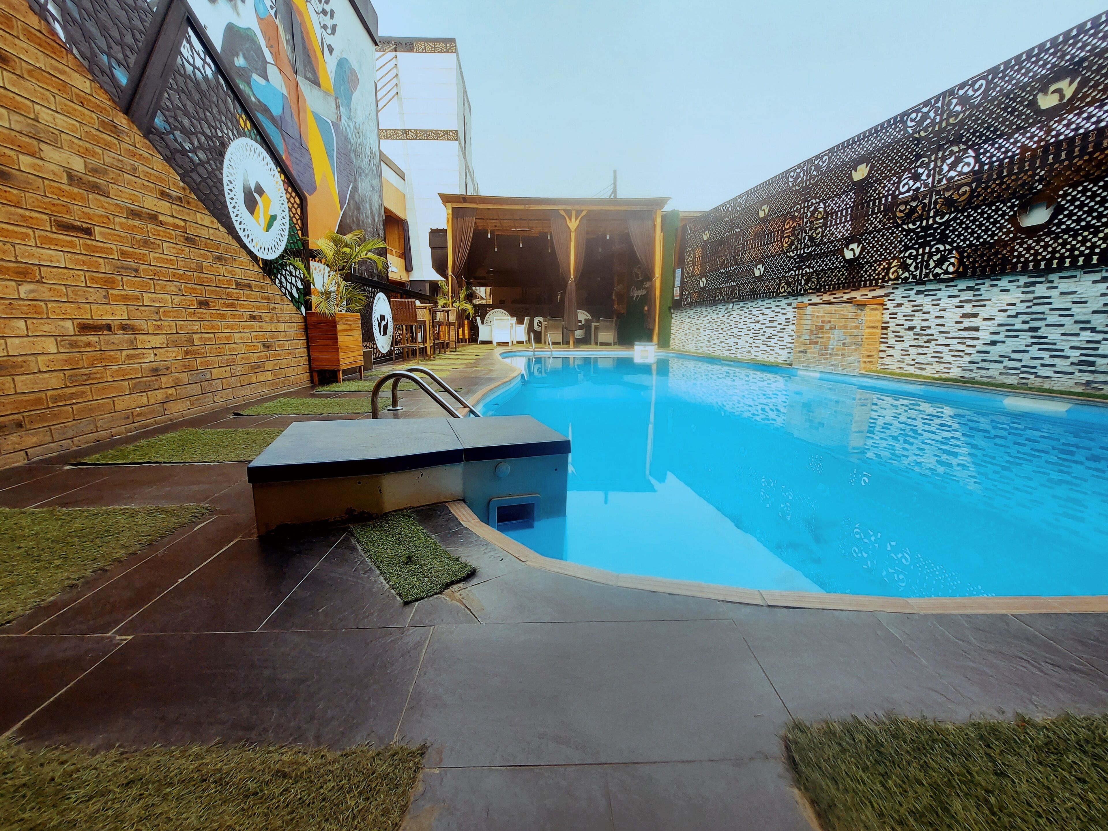 A swimming pool with a tiled deck, a covered seating area, and a brick wall with a mural.
