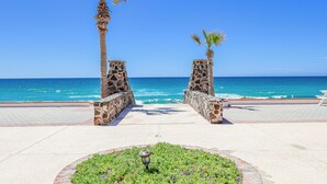 A paved walkway with stone pillars and palm trees, leading to a beach with clear blue water.