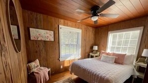 Une chambre avec des murs en bois, un lit avec une literie blanche, un ventilateur de plafond et une fenêtre avec des stores.