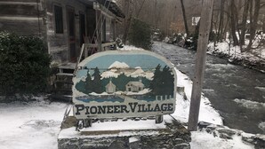 Un panneau indiquant l’entrée du « Village des pionniers », avec une maison en bois rustique et une rivière en arrière-plan.