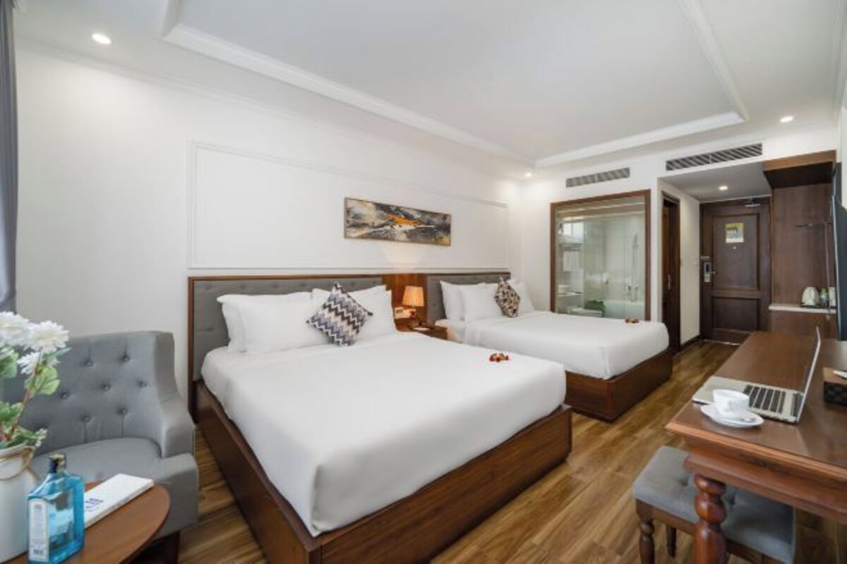 A hotel room with two beds, a desk with a laptop, a chair, and a small table with a vase and bottle.