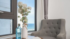 Une pièce avec vue sur l’océan, un fauteuil gris, une petite table ronde avec une bouteille et un livre.