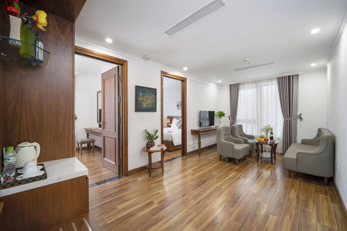 Ein modernes Hotelzimmer mit Holzboden, einer Sitzecke mit zwei Sesseln und einem kleinen Tisch, einem Fernseher und einem durch eine offene Tür einsehbaren Badezimmer.