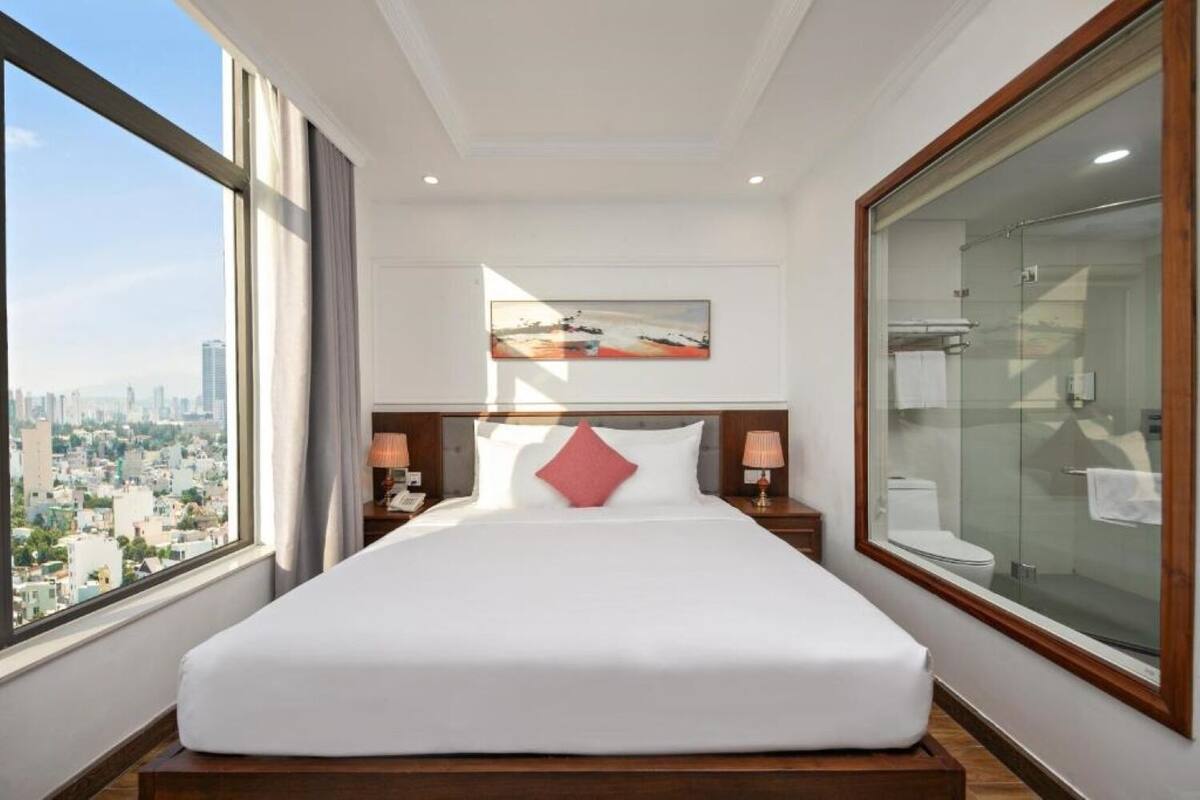 Ein Hotelzimmer mit großem Bett, Blick auf die Stadt, einem Gemälde an der Wand und einer Duschkabine aus Glas.