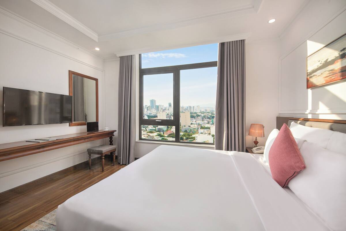 Ein Hotelzimmer mit einem großen Bett, einem Schreibtisch, einem Fernseher und einem Fenster mit Blick auf die Stadt.