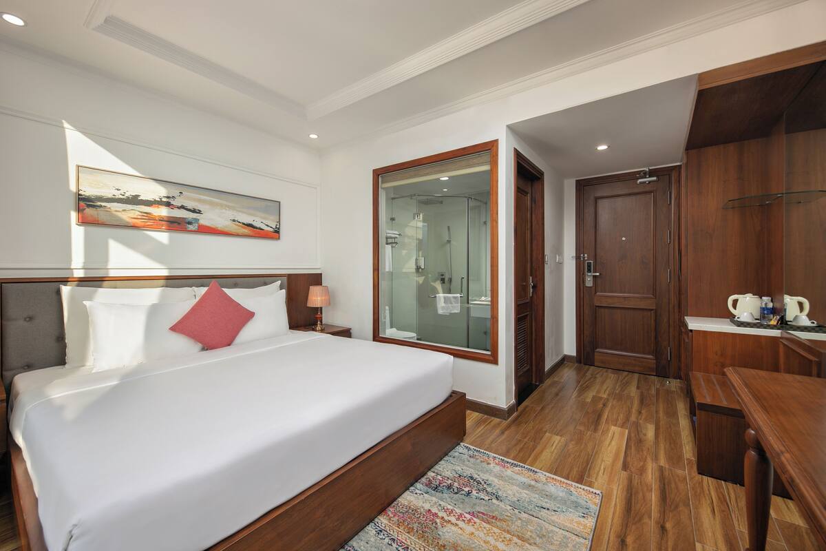 Ein Hotelzimmer mit einem großen Bett, einem Holztisch, einer Duschkabine aus Glas und einem Bild an der Wand.