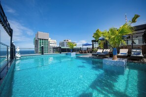 En pool på taget med krystalklart vand, liggestole og udsigt til havet og bygninger.