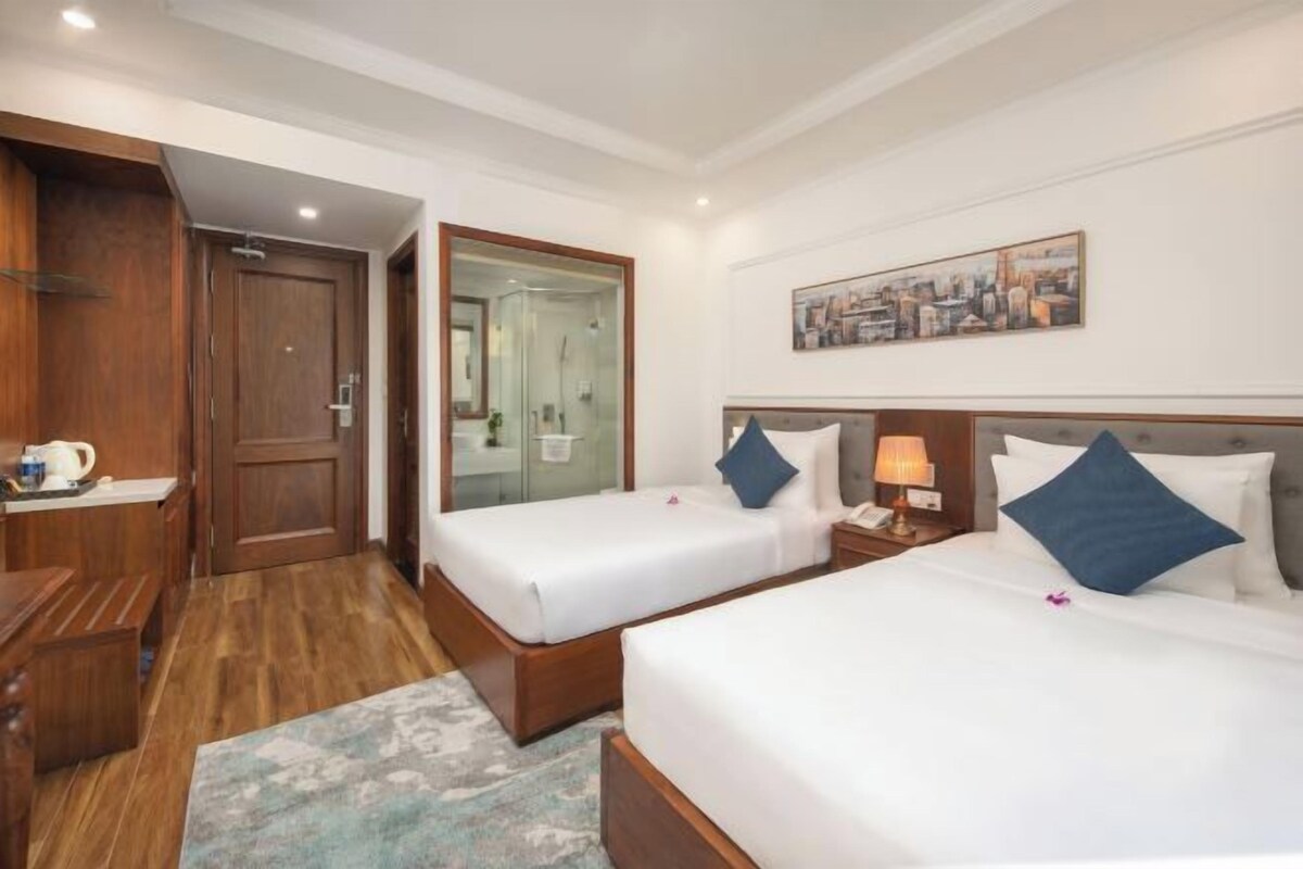 Habitación de hotel con dos camas, un armario de madera, un baño con puerta de vidrio y un cuadro en la pared.
