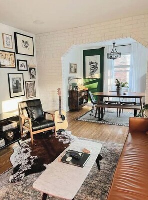 Uma sala de estar aconchegante com poltrona de couro, mesa de centro, um violão e um espaço de jantar com mesa e cadeiras.